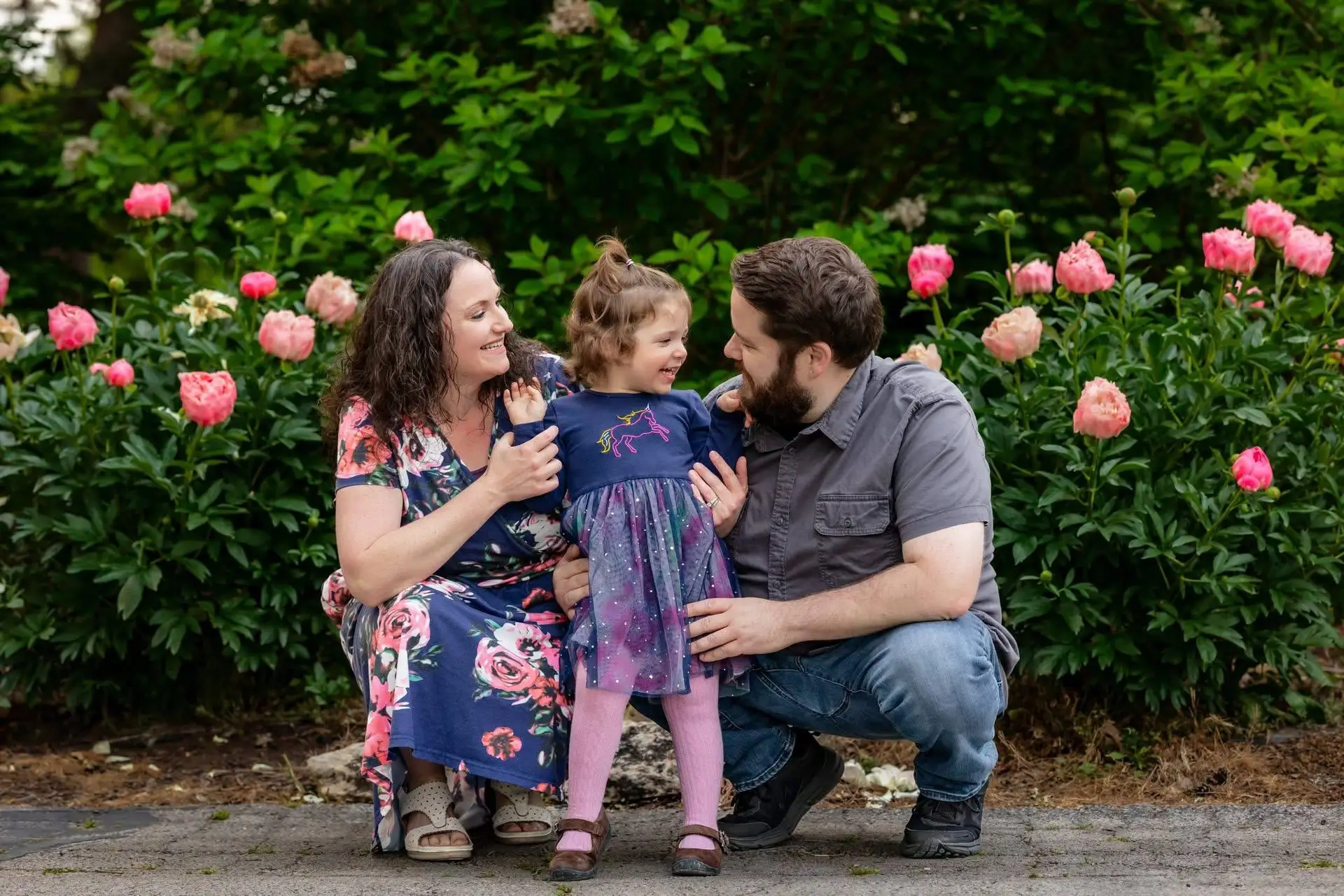 A woman, a man, and a young girl smile and laugh together in front of blooming pink flowers, framed perfectly by a wedding photographer. The woman wears a floral dress, the man is in gray, and the girl shines in a blue dress. A woman, a man, and a young girl smile and laugh together in front of blooming pink flowers, framed perfectly by a wedding photographer. The woman wears a floral dress, the man is in gray, and the girl shines in a blue dress.