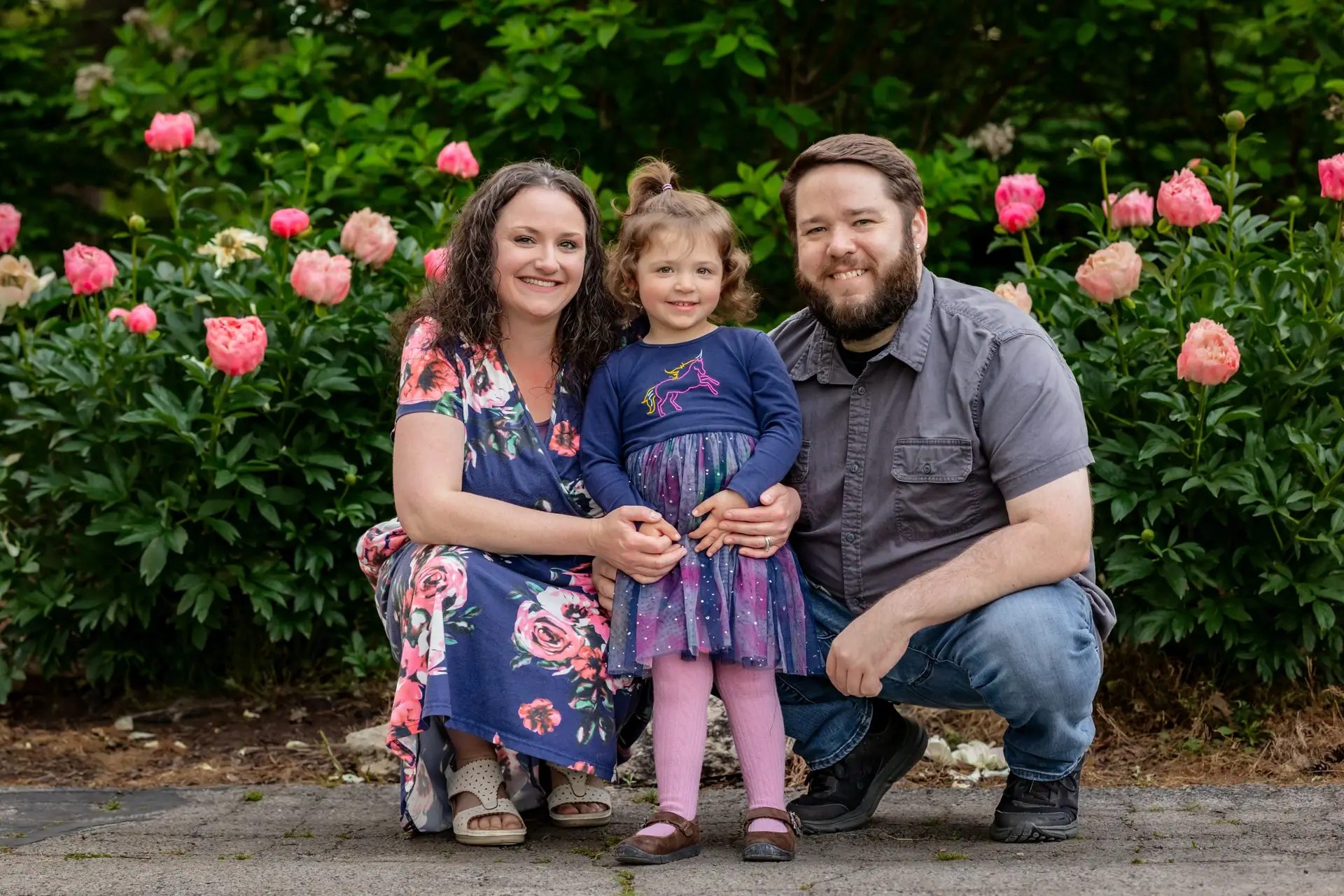 A smiling family of three—a woman in a floral dress, a young girl in a blue dress, and a man in a gray shirt—pose together outside in front of green bushes with pink flowers.