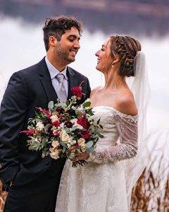 A bride in a white lace dress and veil holds a bouquet of flowers and smiles at a groom in a black suit and tie. They stand outdoors near water, looking happily at each other.