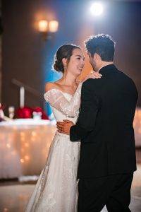 A bride and groom share a joyful first dance at their wedding reception, gazing at each other and smiling. The bride wears a white lace dress, and the groom is in a black suit. Warm lights and a decorated table are in the background.