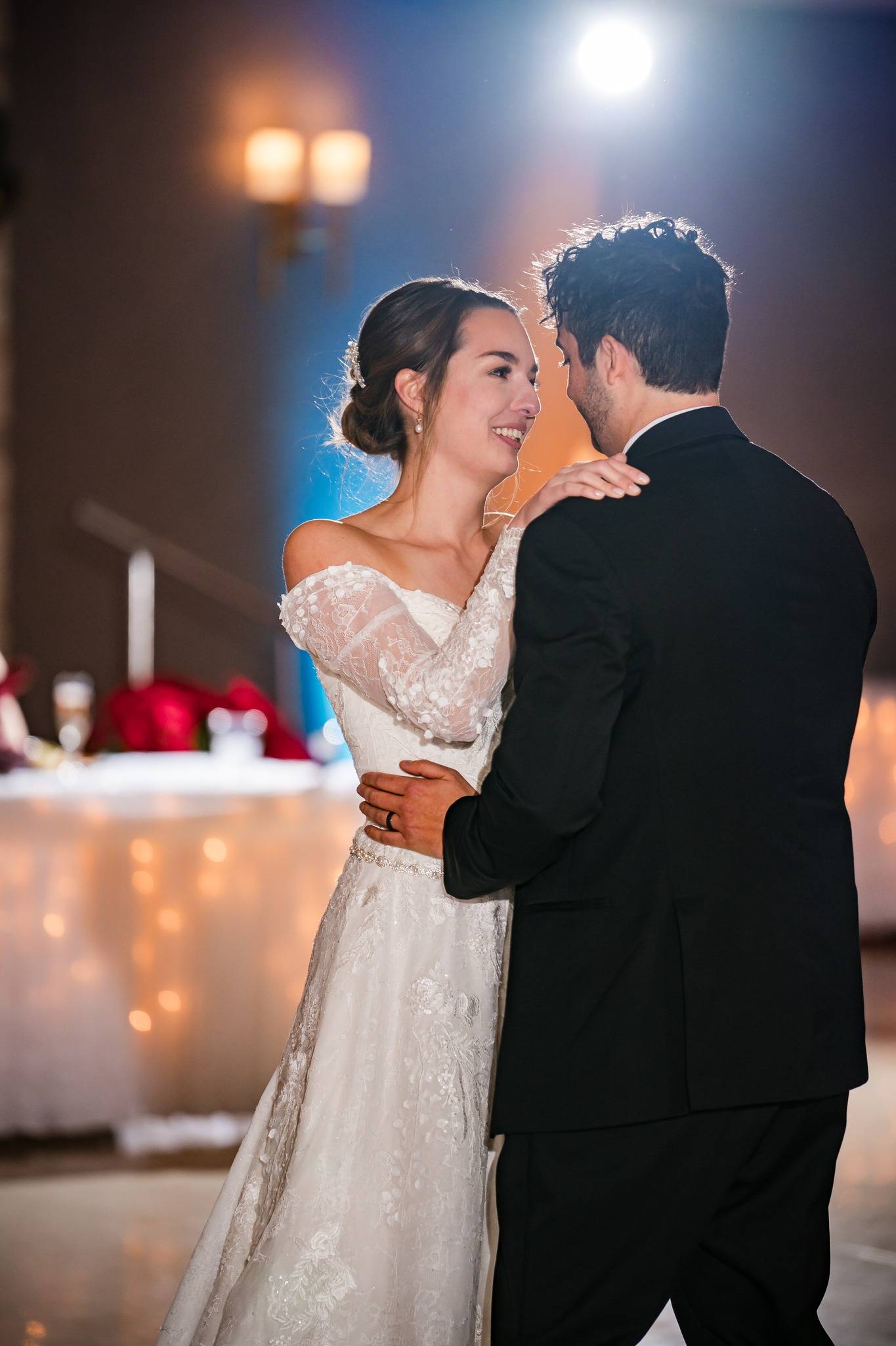 A bride and groom share a joyful first dance at their wedding reception, gazing at each other and smiling. The bride wears a white lace dress, and the groom is in a black suit. Warm lights and a decorated table are in the background.