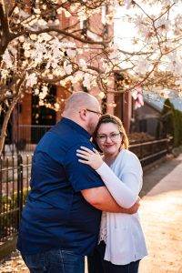 A couple embraces under a flowering tree on a sunlit sidewalk; the man kisses the womans forehead while she smiles at the camera, wearing glasses and a white cardigan.