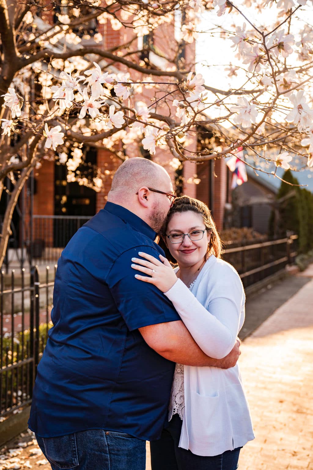 A couple embraces under a flowering tree on a sunlit sidewalk; the man kisses the womans forehead while she smiles at the camera, wearing glasses and a white cardigan.