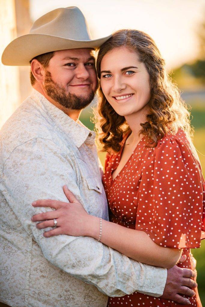 A smiling couple embraces outdoors at sunset. The man wears a cowboy hat and patterned shirt, while the woman, in a red polka dot dress, rests her hand on his chest, her engagement ring sparkling—perfect for wedding photography packages.