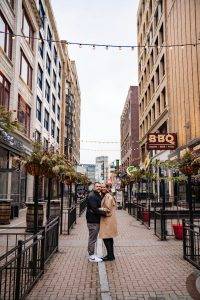 A couple stands close together, smiling and hugging in the middle of a pedestrian street lined with restaurants and lights, with tall buildings on both sides.