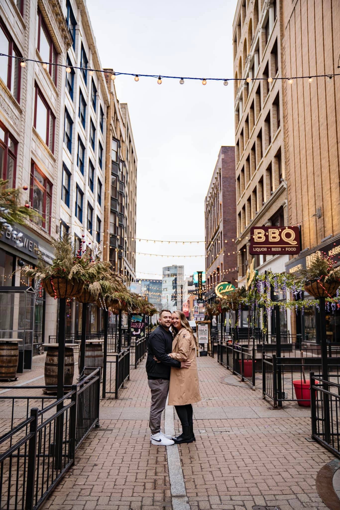 A couple stands close together, smiling and hugging in the middle of a pedestrian street lined with restaurants and lights, with tall buildings on both sides.