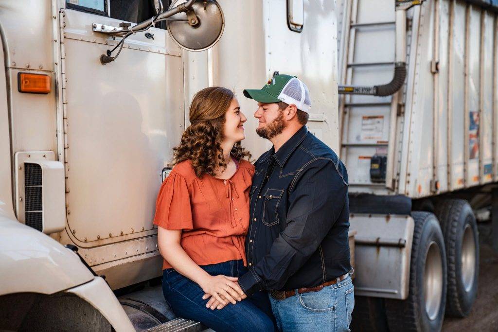 A couple sits on the step of a large white truck, holding hands and smiling at each other—capturing a candid moment perfect for wedding photography packages. The woman wears an orange top; the man wears a black shirt, jeans, and a cap.