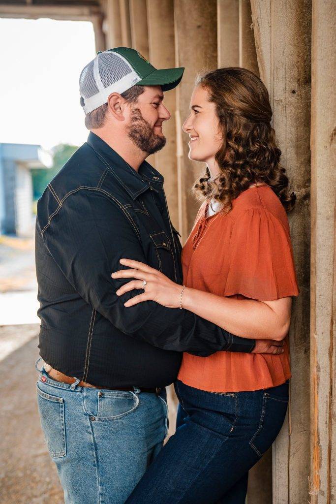 A couple stands close together facing each other, smiling, with the womans arms around the mans shoulders. Outdoors near a wooden structure in casual clothing, they look happy—perfect for couples exploring wedding photography packages.