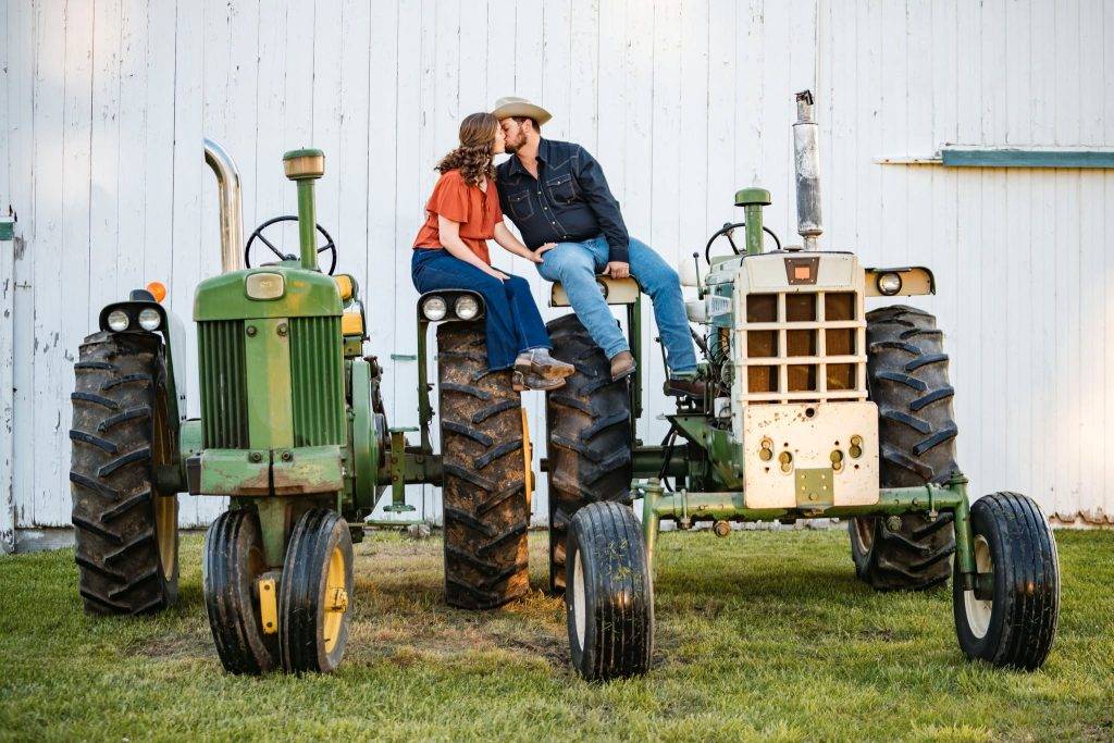 A couple sits on separate vintage tractors, leaning in for a kiss, captured perfectly for wedding photography packages. She wears a red blouse and jeans; he sports a cowboy hat. They pose before a white wooden barn wall.