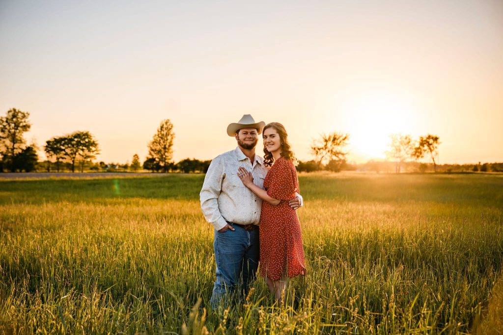 A couple stands together in a field at sunset. The man wears a cowboy hat, glasses, a light shirt, and jeans. The woman wears a red dress with white polka dots. Both smile and embrace with sunlight—perfect for wedding photography packages.