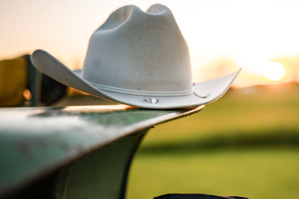 A light gray cowboy hat rests on the edge of a green metal surface, with a blurred field and setting sun in the background—a perfect scene often captured in rustic wedding photography packages.