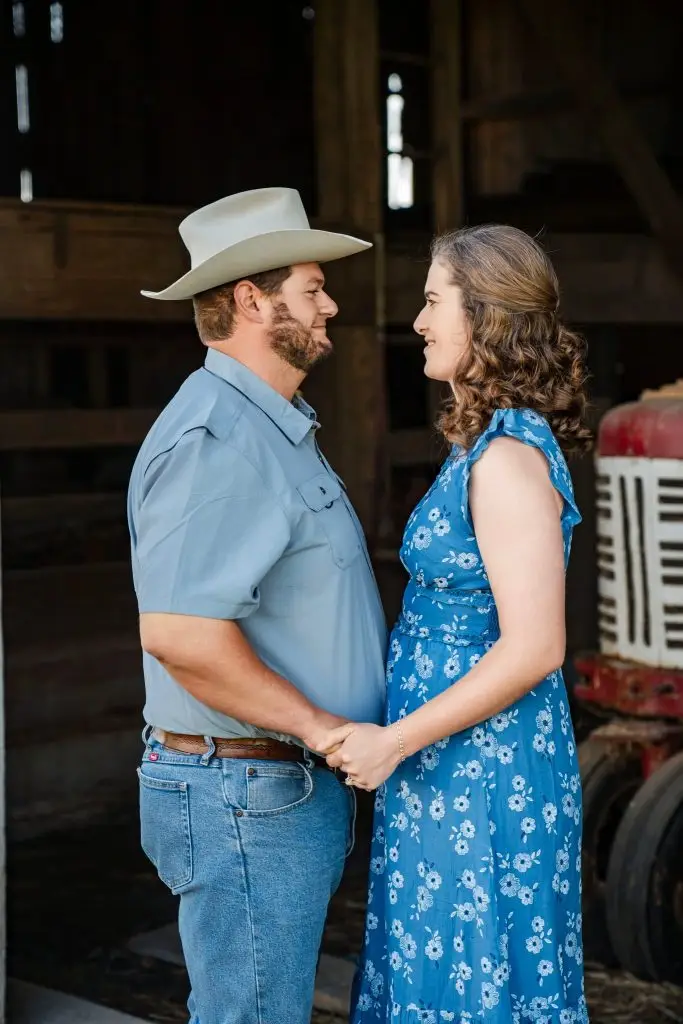 A man in a cowboy hat and a woman in a blue floral dress stand facing each other, holding hands and smiling, in front of a rustic barn and an old red tractor—perfect for unique wedding photography packages.