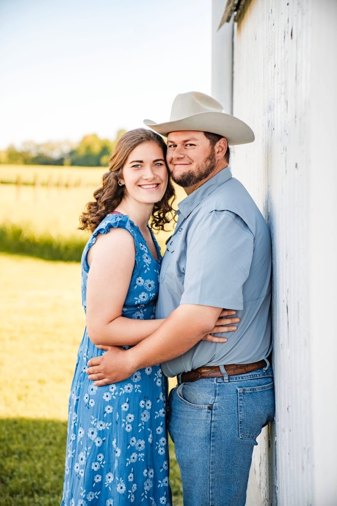 A smiling couple stands close together by a white barn wall in a sunny field. Perfect for showcasing wedding photography packages, the woman wears a blue floral dress and the man, in a cowboy hat and jeans, embraces her as they look at the camera.