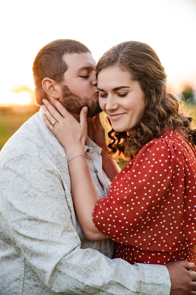 A man kisses a woman on the cheek while gently holding her face. The woman, in a red polka-dot dress, smiles with her eyes closed, savoring the moment outdoors at sunset—perfect inspiration for wedding photography packages.