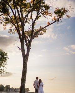 A couple stands side by side under a tall tree, looking out at a scenic view during sunset. The sky is partly cloudy with soft sunlight, and houses are visible in the distance.