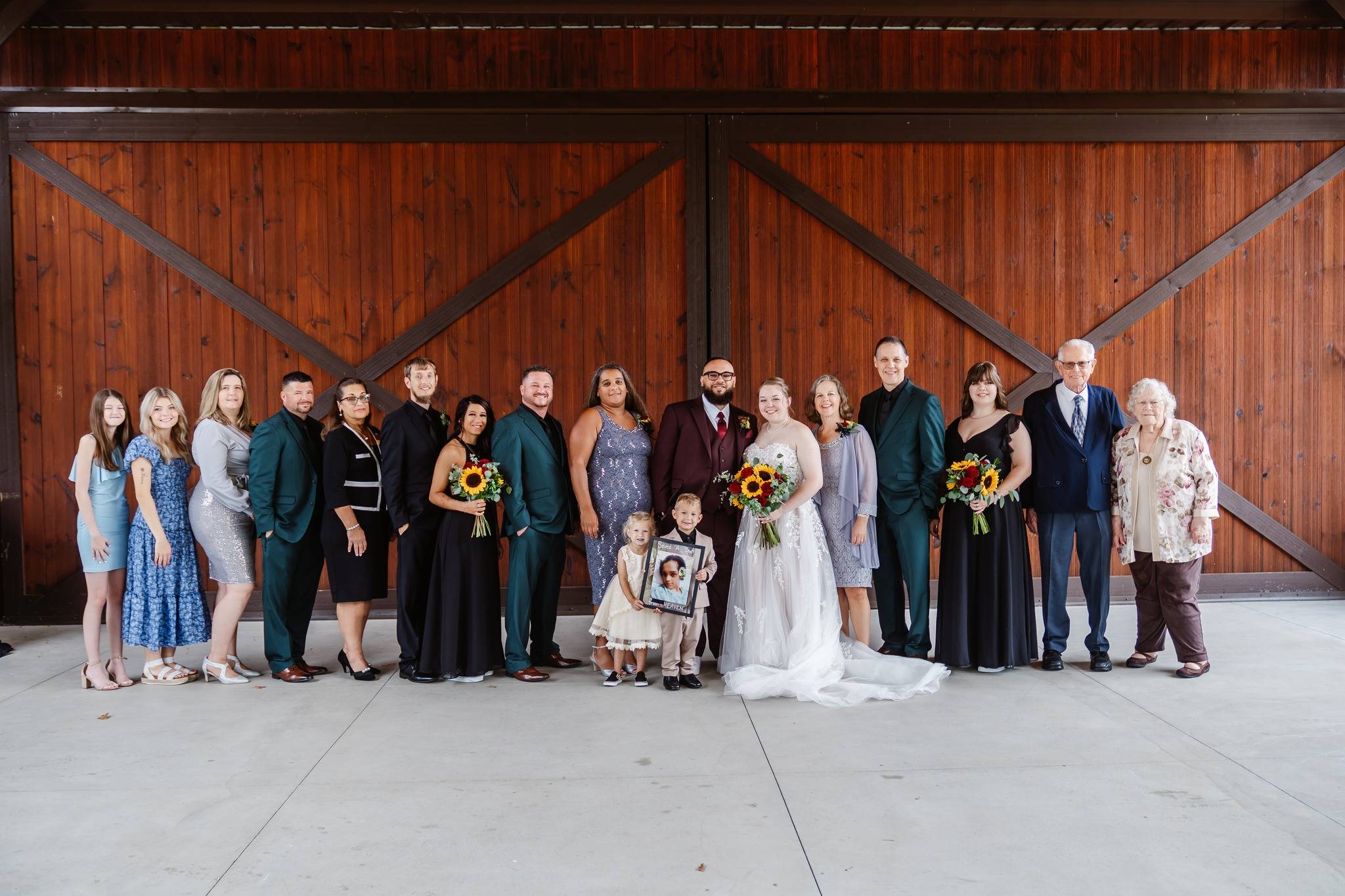 A wedding party poses in front of large wooden doors. The bride and groom stand in the center holding flowers, surrounded by family and friends, including children in formal attire. Everyone is smiling.