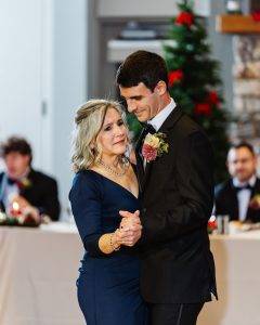 A woman in a navy blue dress and a man in a black suit share a dance at a formal event. They look emotional and close. A decorated Christmas tree and people seated at a table are visible in the background.