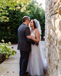 A bride and groom stand close, smiling and touching noses in an outdoor garden setting. The bride wears a white gown and veil, while the groom is in a gray suit. Sunlight filters through greenery and stone walls around them.