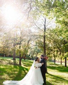 A bride and groom embrace in a sunlit park surrounded by green trees. The bride wears a long white dress with a veil, holding a bouquet, while sunlight streams through the branches above.