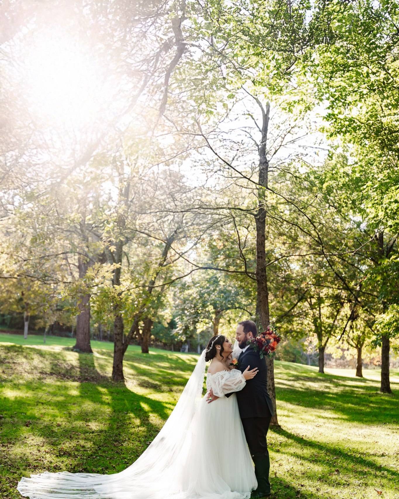 A bride and groom embrace in a sunlit park surrounded by green trees. The bride wears a long white dress with a veil, holding a bouquet, while sunlight streams through the branches above.