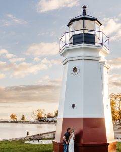 A couple embraces at the base of a white and red lighthouse by the water, with trees, a bench, and a partly cloudy sky in the background at sunset.