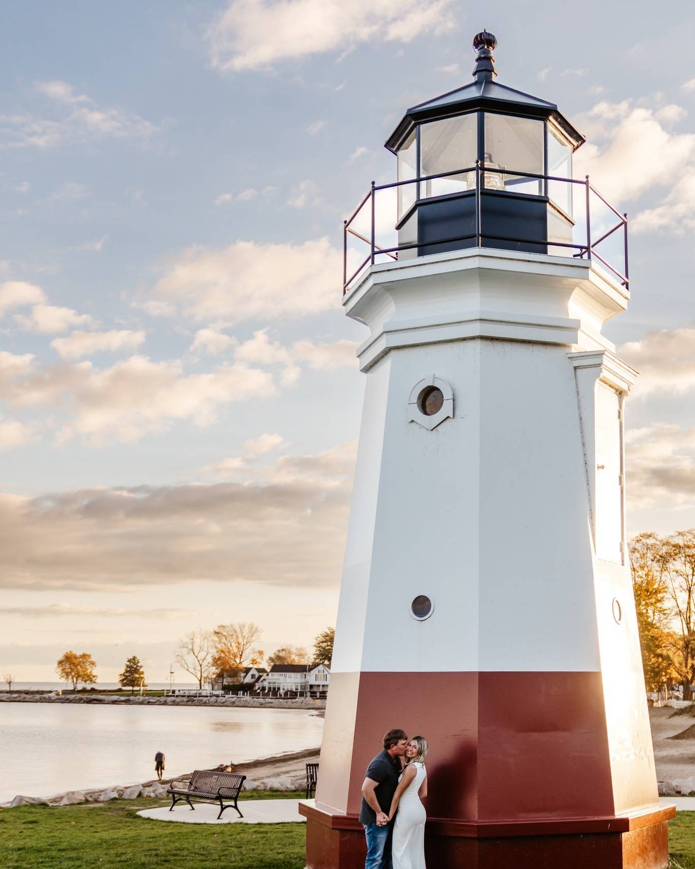 A couple embraces at the base of a white and red lighthouse by the water, with trees, a bench, and a partly cloudy sky in the background at sunset.