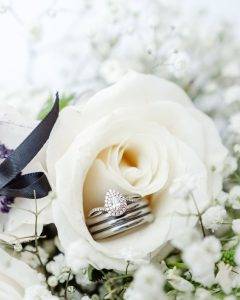 A close-up of a diamond engagement ring and matching wedding bands nestled in the center of a white rose, surrounded by babys breath and a black ribbon.