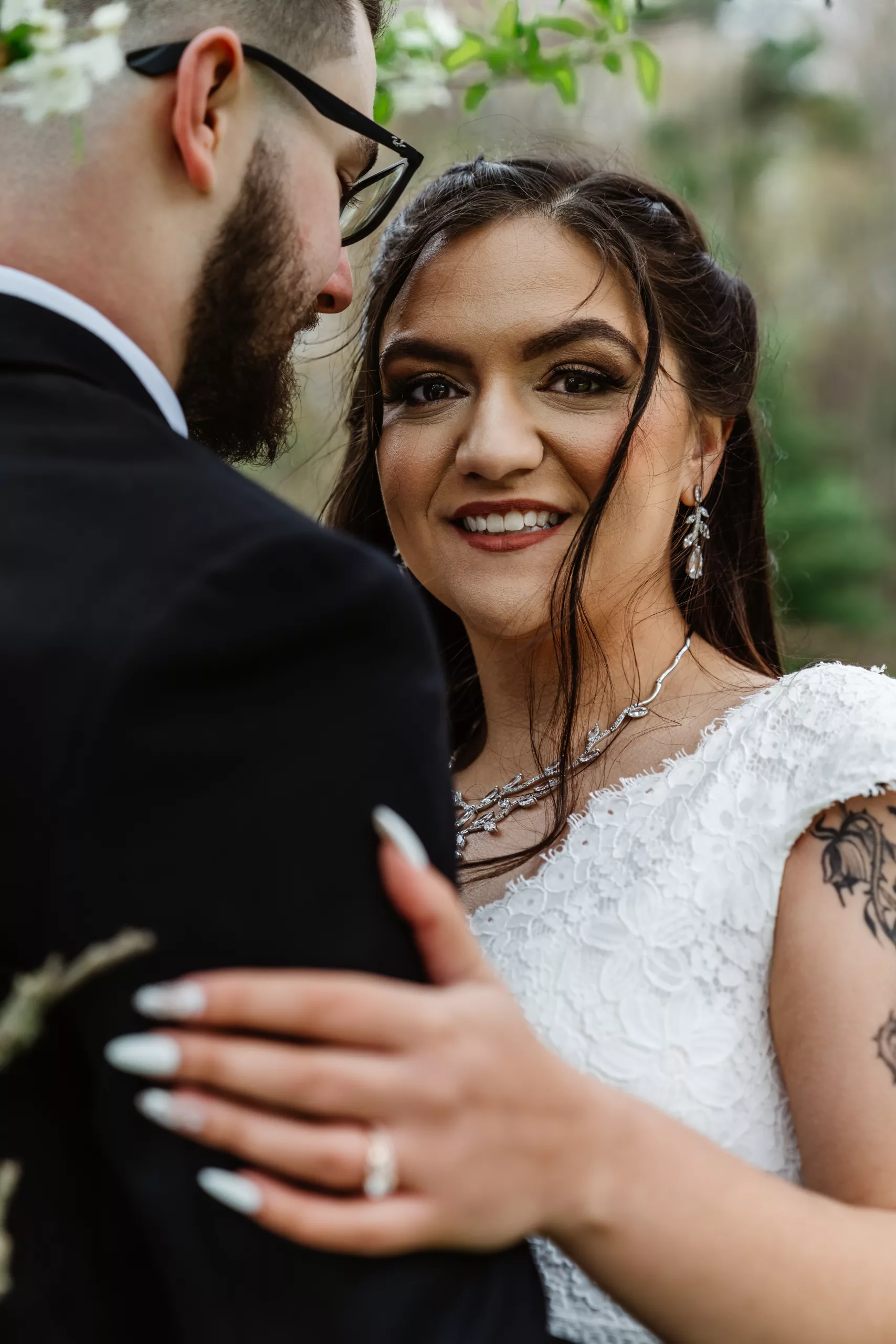 A bride in a white lace dress smiles at the camera while embracing a groom in a black suit. She has long nails, a visible tattoo, and wears elegant jewelry. Greenery is blurred in the background. A bride in a white lace dress smiles at the camera while embracing a groom in a black suit. She has long nails, a visible tattoo, and wears elegant jewelry. Greenery is blurred in the background.