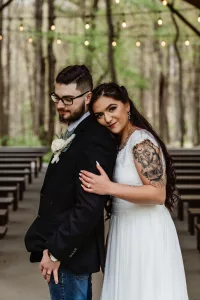 A bride in a white dress leans affectionately on a groom in a suit and jeans. They stand under string lights in an outdoor, wooded venue with empty benches behind them.