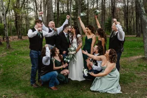 A wedding party celebrates outdoors in a forested area. The bride and groom kiss in the center while bridesmaids and groomsmen around them cheer, smiling and raising bouquets and arms in excitement.
