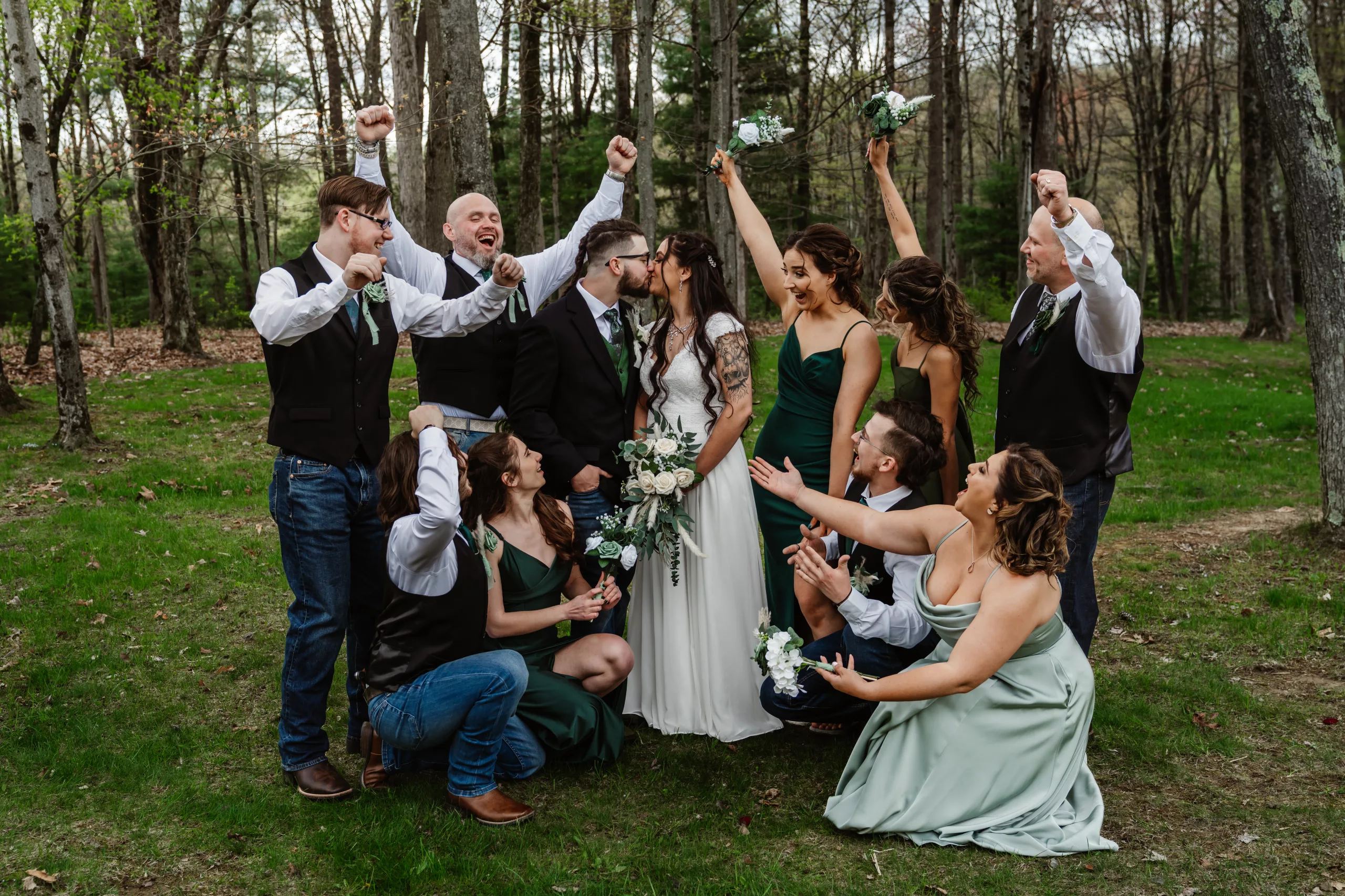 A wedding party celebrates outdoors in a forested area. The bride and groom kiss in the center while bridesmaids and groomsmen around them cheer, smiling and raising bouquets and arms in excitement.