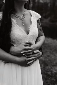 A black and white photo of a bride in a lace wedding dress, standing outdoors, with someone’s arms wrapped around her waist from behind. The bride wears a decorative necklace and rings, with long hair and visible tattoos.