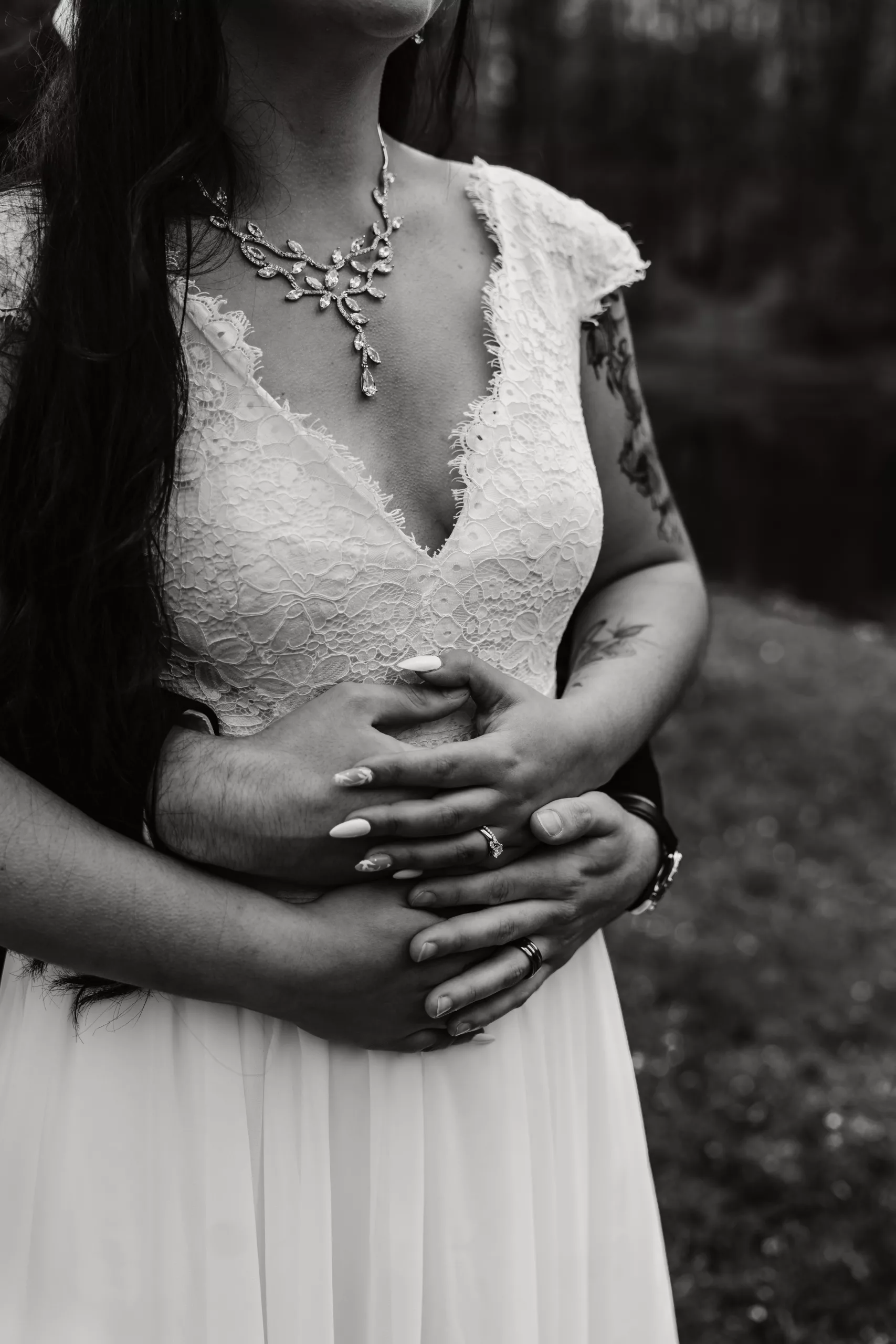 A black and white photo of a bride in a lace wedding dress, standing outdoors, with someone’s arms wrapped around her waist from behind. The bride wears a decorative necklace and rings, with long hair and visible tattoos.