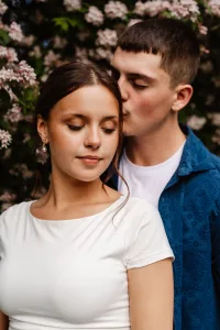 A young man gently kisses a young woman on the temple as she stands in front of him, eyes closed and smiling softly, with blooming flowers in the background.