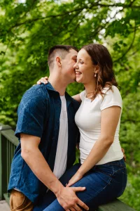 A young couple smiles as the man whispers in the womans ear outdoors. They sit close together on a wooden railing surrounded by lush green trees, with the woman wearing a white shirt and jeans and the man in a blue shirt.