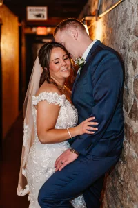 A bride in a lace wedding dress and veil smiles with her eyes closed as a groom in a blue suit gently rests his forehead on hers, standing close together against a stone wall in a warmly lit hallway.