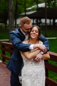 A groom in a blue suit hugs his smiling bride in a white lace wedding dress from behind as they stand on a wooden bridge outdoors, surrounded by greenery and trees.