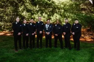 Eight men dressed in black suits with boutonnieres stand in a row on grass in front of tall pine trees, posing for a group photo outdoors.
