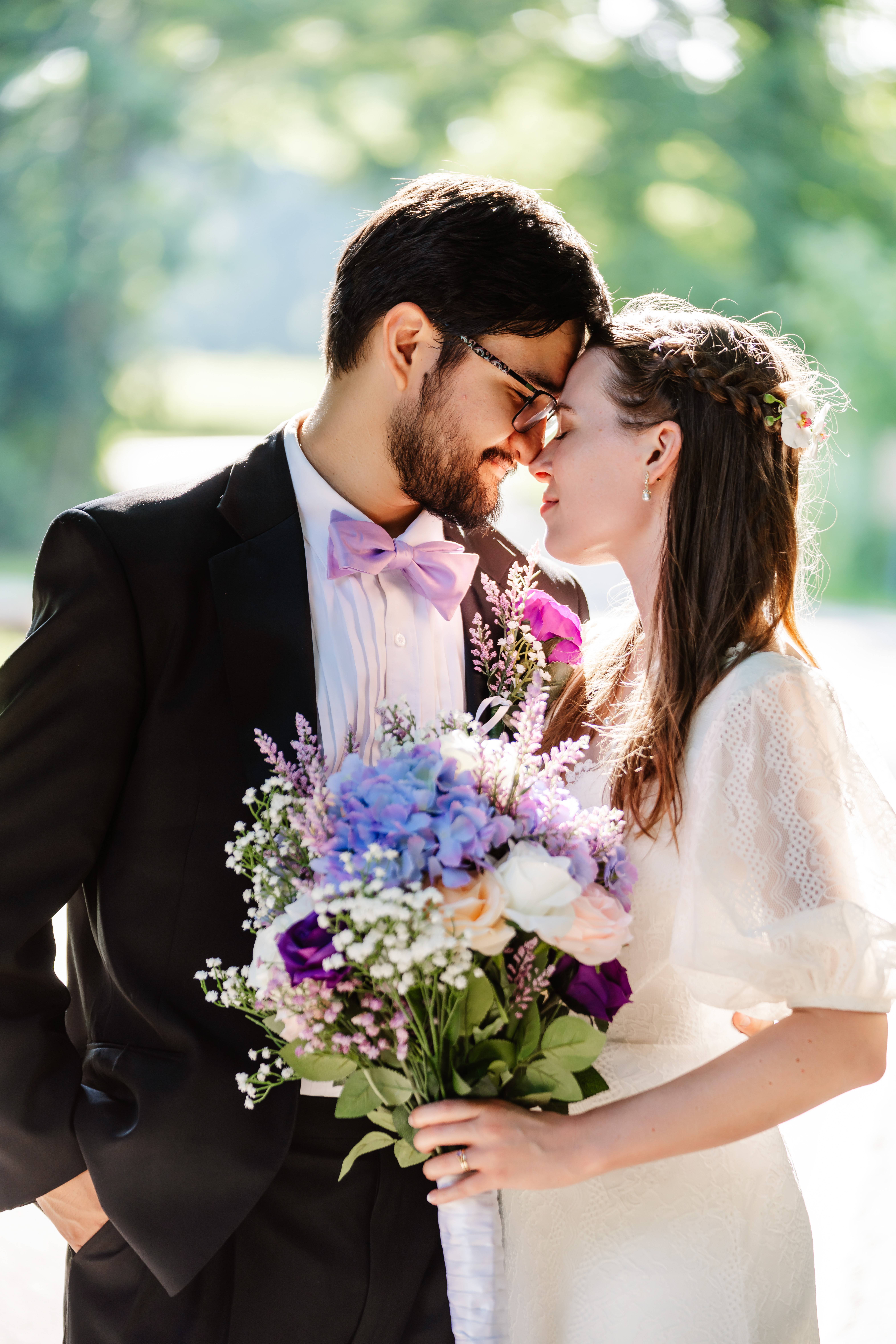 A bride and groom stand close together, smiling with their foreheads touching. Captured by a wedding photographer, the bride holds a bouquet of purple, white, and lavender flowers as sunlight filters through trees in the background. A bride and groom stand close together, smiling with their foreheads touching. Captured by a wedding photographer, the bride holds a bouquet of purple, white, and lavender flowers as sunlight filters through trees in the background.