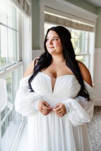 A woman in an off-the-shoulder white wedding dress stands by a window, looking outside with a soft smile. A wedding photographer captures her long dark hair and natural makeup as sunlight gently illuminates her face.
