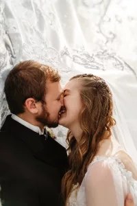 A bride and groom gently touch noses and smile under a lace veil, sharing an intimate, joyful moment on their wedding day. Soft sunlight filters through the delicate fabric above them.