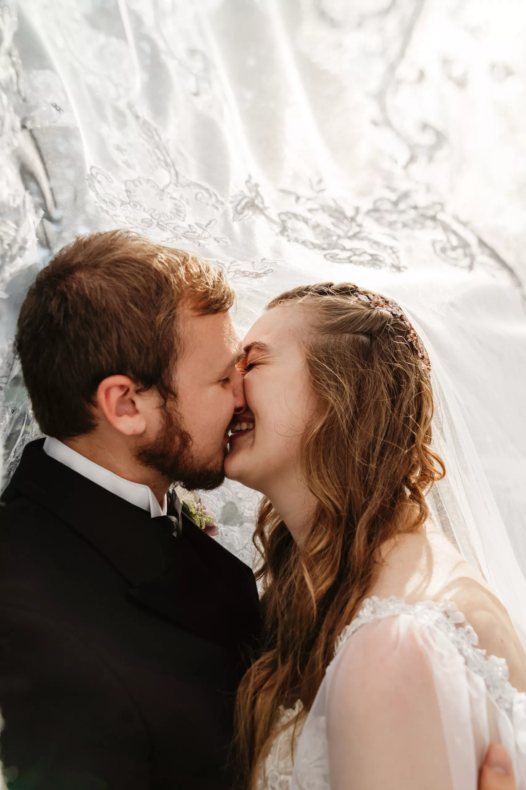 A bride and groom gently touch noses and smile under a lace veil, sharing an intimate, joyful moment on their wedding day. Soft sunlight filters through the delicate fabric above them.