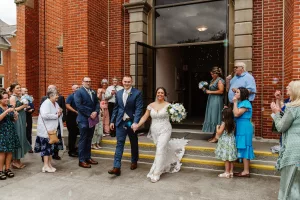 A bride and groom walk hand in hand out of a brick building, smiling as wedding guests stand on either side, clapping and blowing bubbles to celebrate their marriage. The bride holds a bouquet of white flowers.