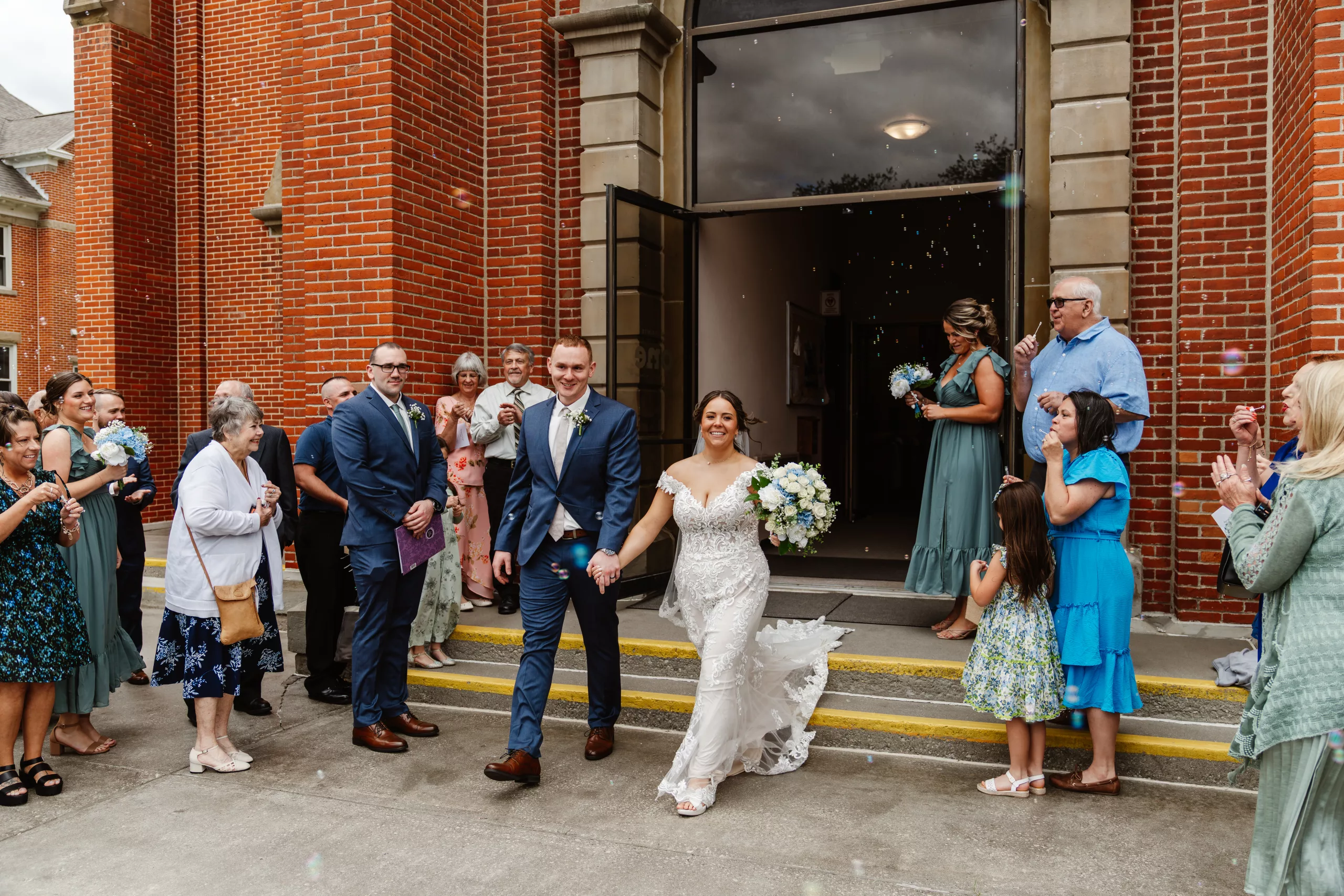 A bride and groom walk hand in hand out of a brick building, smiling as wedding guests stand on either side, clapping and blowing bubbles to celebrate their marriage. The bride holds a bouquet of white flowers.