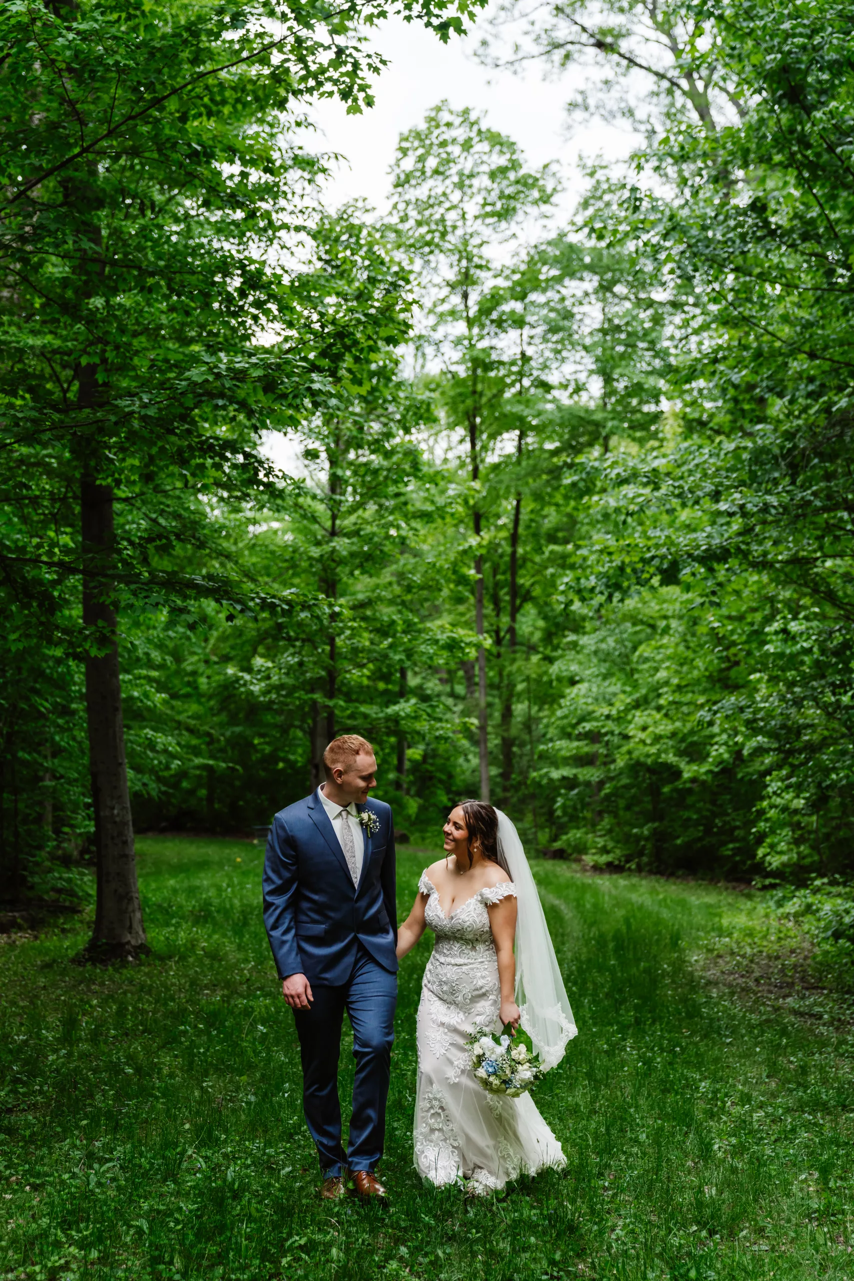 A bride and groom walk hand in hand through a lush, green forest. The bride wears a white dress and veil, holding a bouquet, while the groom is in a blue suit. Both are smiling, surrounded by tall trees and grass.