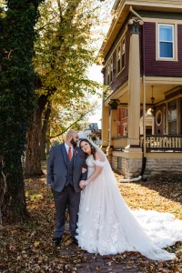 A bride in a white gown leans on the groom, who wears a gray suit and orange tie, as their wedding photographer captures them standing on a leaf-covered sidewalk by a large tree and historic house in autumn daylight.