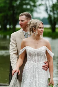 A bride in an off-the-shoulder white lace dress stands on a wooden bridge, holding a bouquet, while a groom in a beige suit stands behind her with his arm around her waist. Green trees and water are in the background.