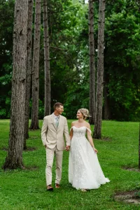 A bride and groom walk hand in hand through a green, wooded area. The bride wears an off-the-shoulder white gown, while the groom is in a light beige suit. Both are smiling and looking at each other.